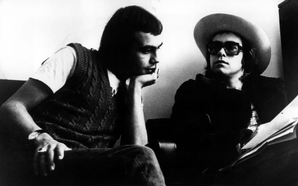 Bernie Taupin, chin on hand, beside Elton John in a wide-brimmed hat and dark glasses holding sheets of paper