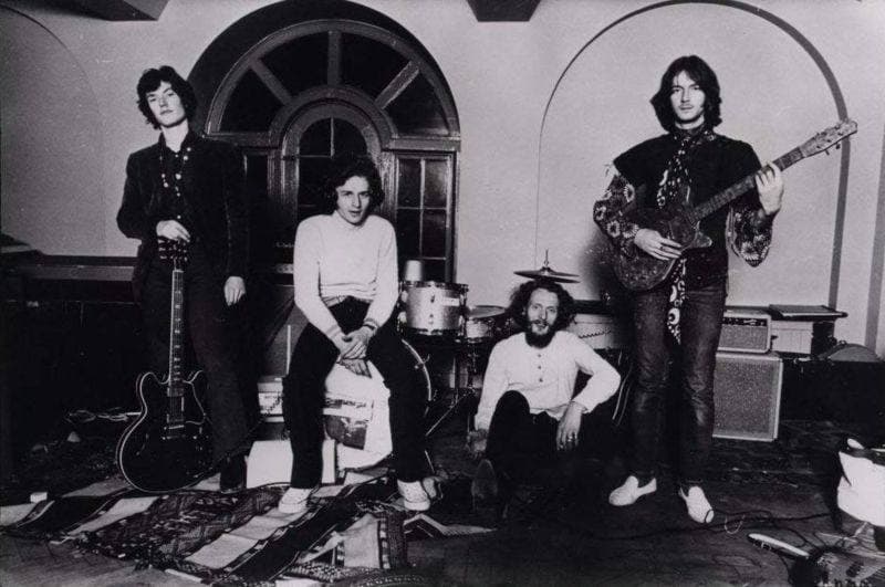 Four musicians with guitars and a drum kit on a rug in a room with arched windows