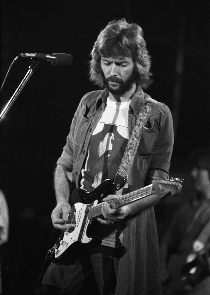 Eric Clapton on stage in a long coat, head bowed over an electric guitar at a microphone