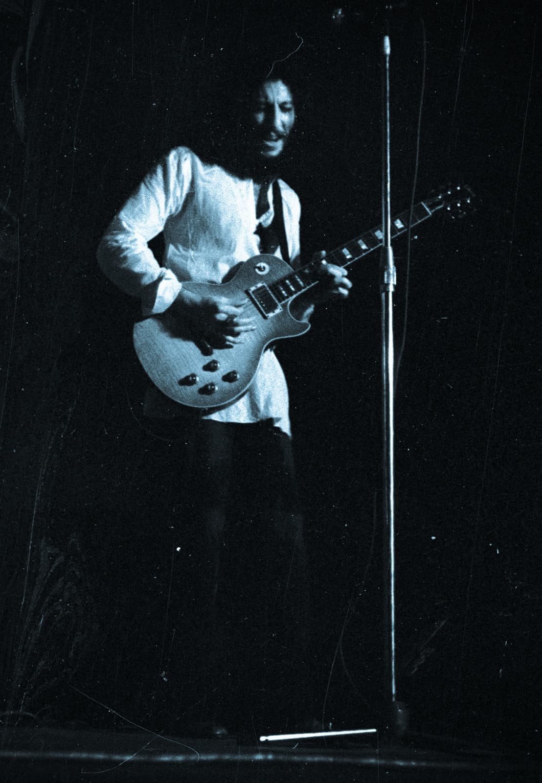 Peter Green in a white shirt playing a Les Paul guitar at a microphone on a dark stage