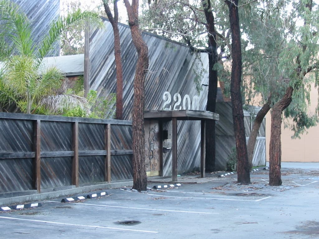 A weathered wooden building front numbered 2200 behind a fence and eucalyptus trunks