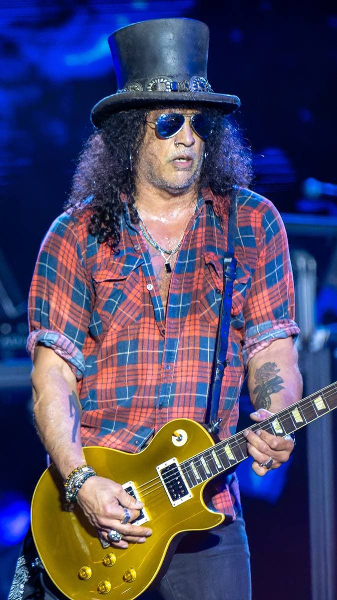 Slash in a top hat and sunglasses playing a guitar on stage