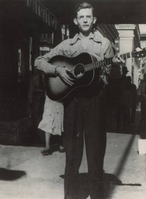 A teenage Hank Williams in a hat playing a guitar on a city pavement