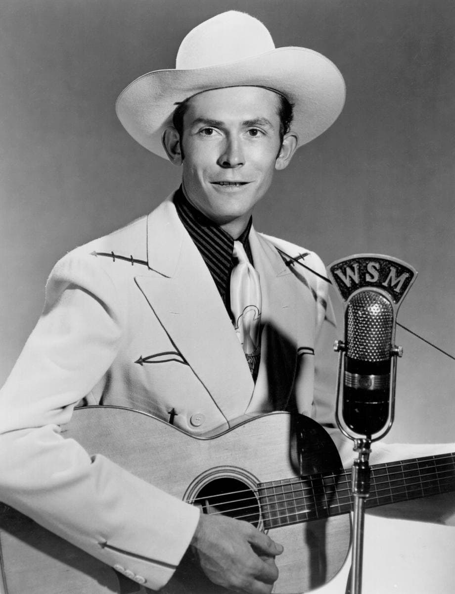 Hank Williams in a white cowboy hat and western suit, holding a guitar and smiling