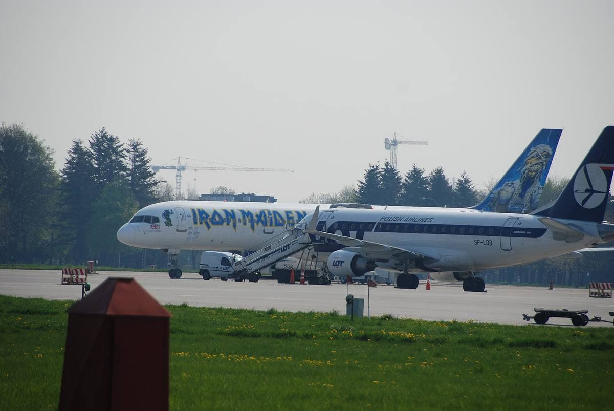 A white airliner with the band name on its fuselage, parked on an apron