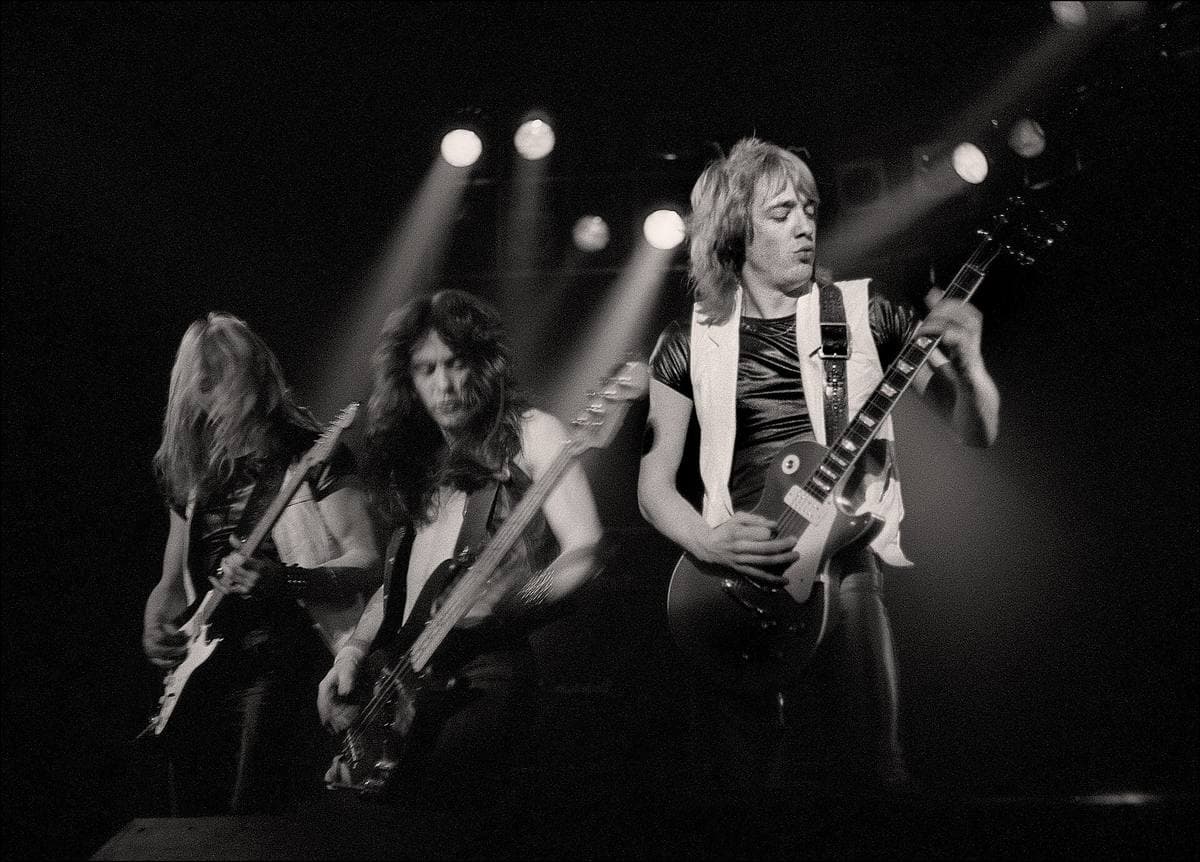 Three guitarists in a row under a rig of lamps, in black and white