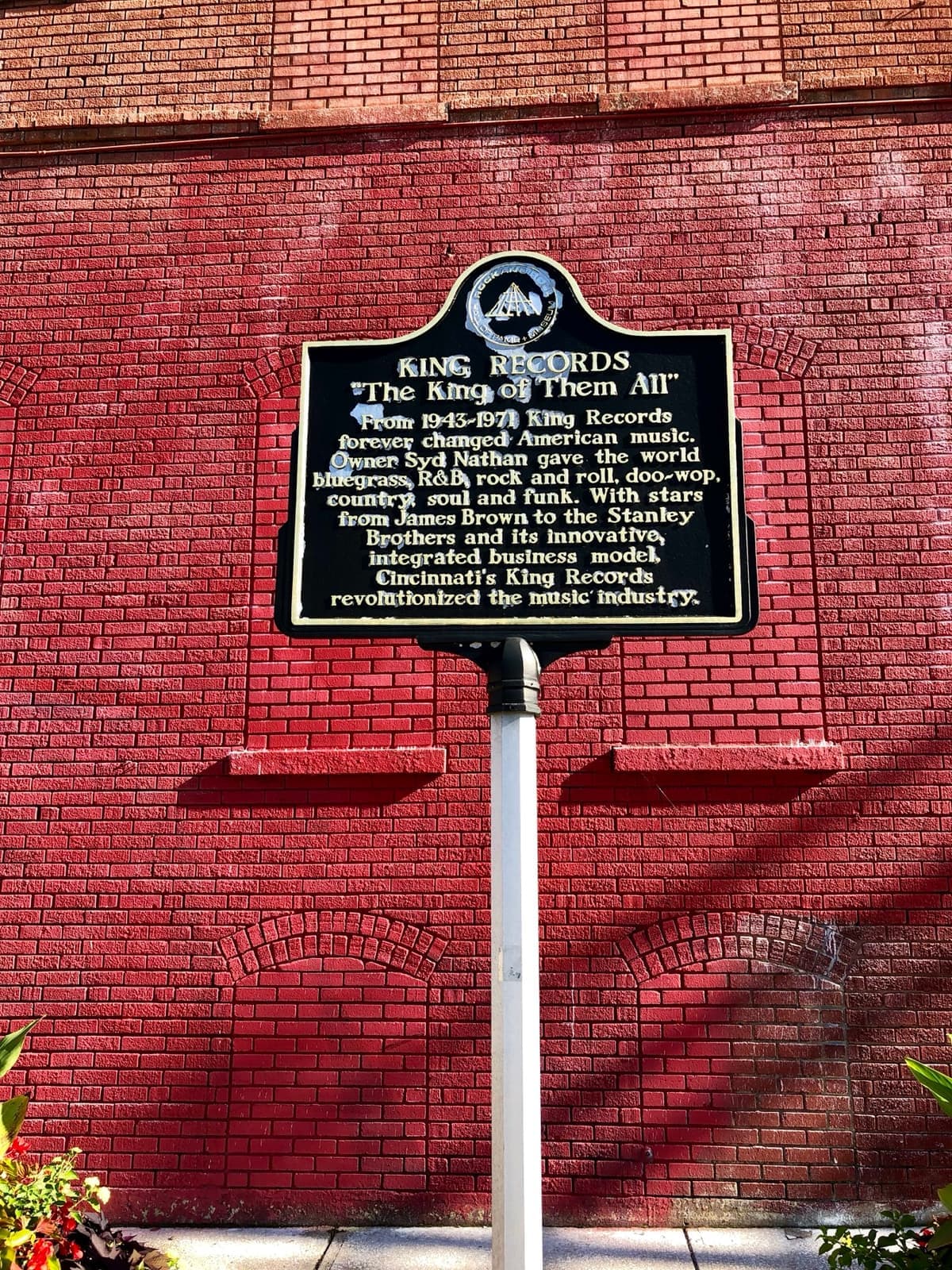 A black historical marker on a pole in front of a red-brick wall