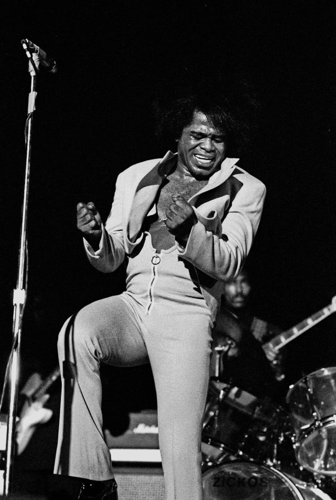 James Brown pointing upwards as he sings, a drum kit behind him