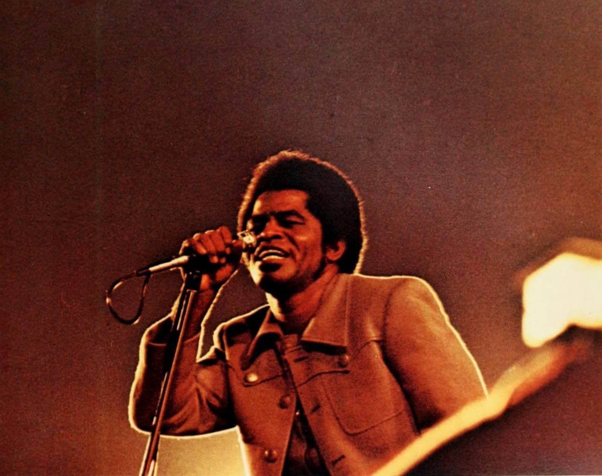 James Brown in a tan suit singing into a microphone in warm orange light