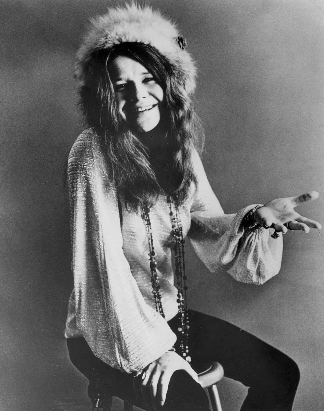 Janis Joplin laughing on a stool in a fur hat and loose blouse, one hand held out with bracelets on the wrist