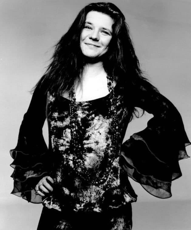 Janis Joplin smiling, hands on hips, in a dark patterned blouse with wide ruffled sleeves