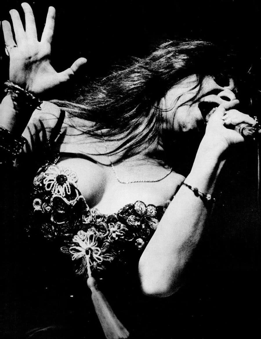 Janis Joplin mid-song, one arm raised, bracelets on both wrists
