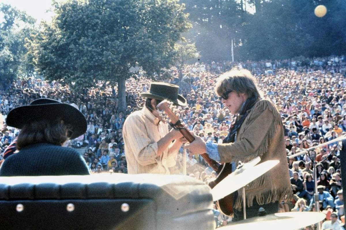 Three musicians in hats and a fringed jacket playing on an outdoor stage in front of a huge crowd under trees