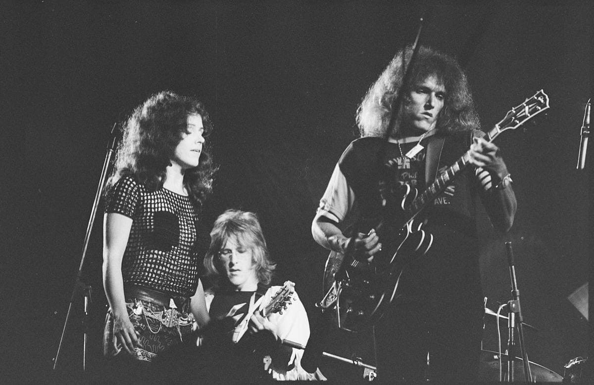 Grace Slick at a microphone beside two guitarists on a dark stage
