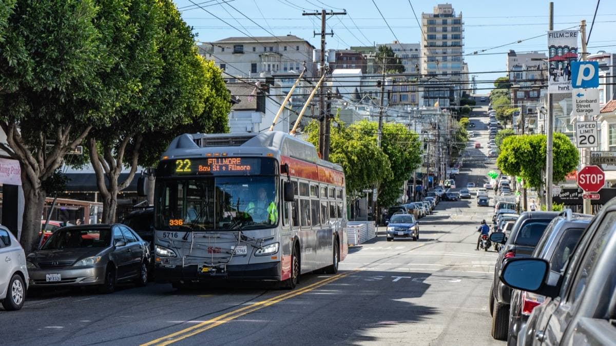 A trolleybus climbing Fillmore Street between parked cars and shopfronts