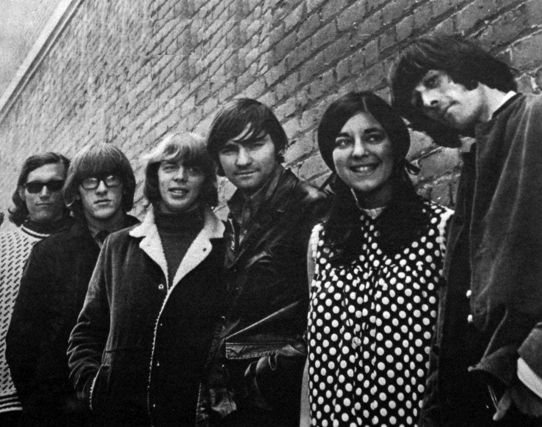 Six band members in dark coats against a brick wall, Signe Anderson in a polka-dot dress
