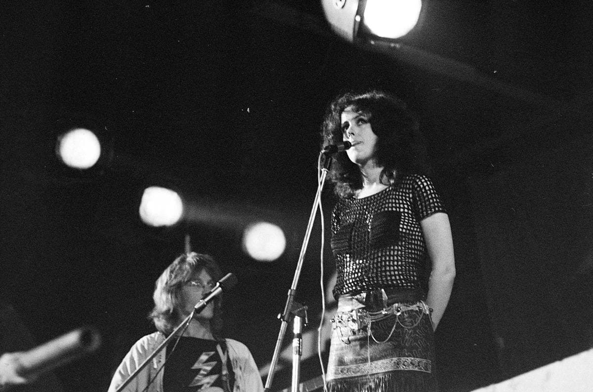 Grace Slick standing at a microphone under three round stage lights, Paul Kantner singing below