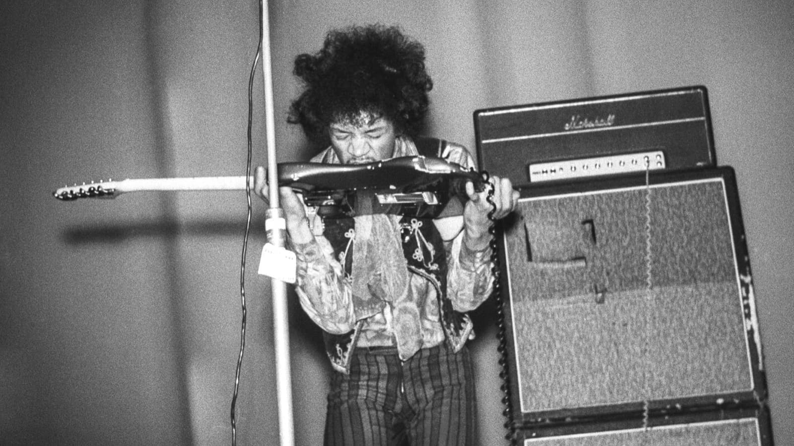 Hendrix lifts his guitar to his mouth beside a stack of amplifiers
