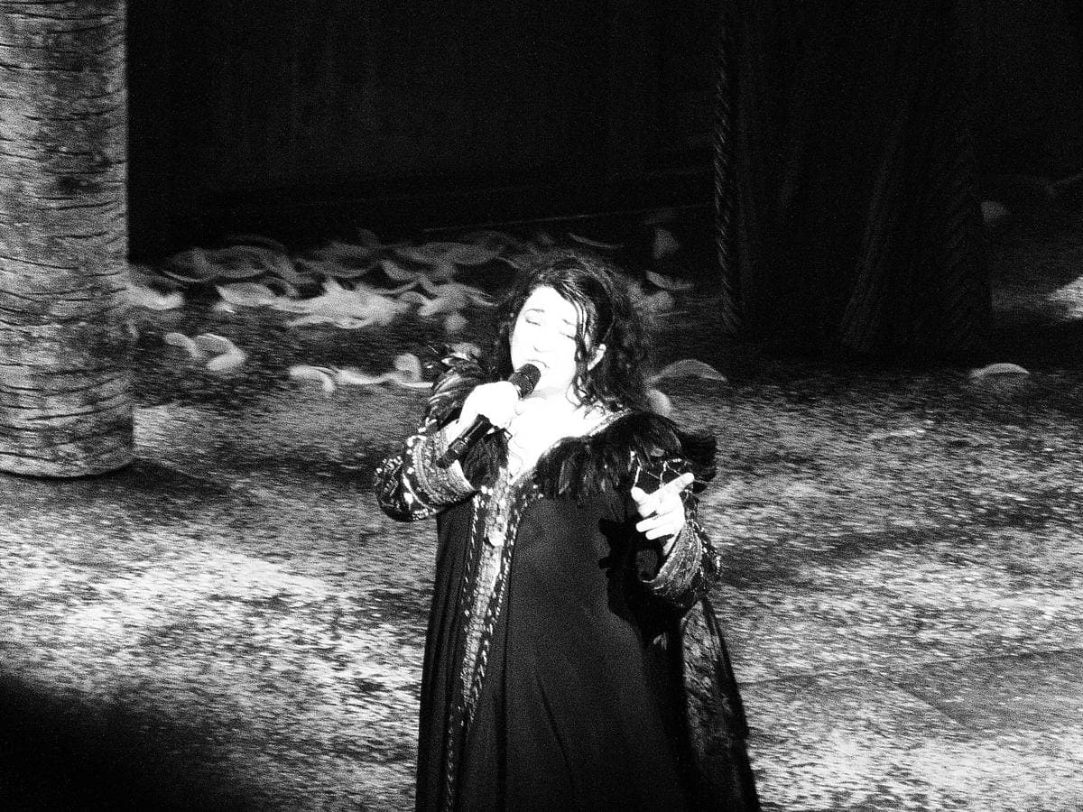 Kate Bush small on a wide stage in a long dark robe, singing into a microphone, a painted backdrop of trees behind her