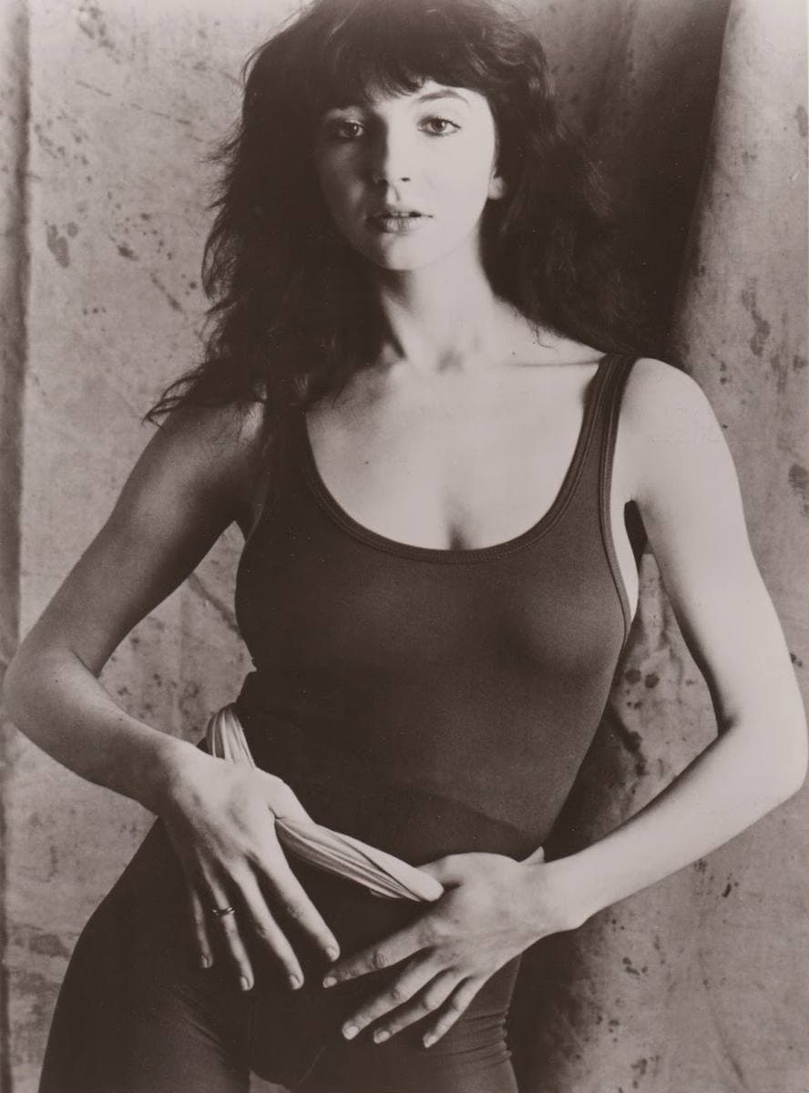 Kate Bush in black and white, aged about nineteen, in a dark leotard, long curly hair over her shoulders, leaning back against a wall with her hands crossed at her waist