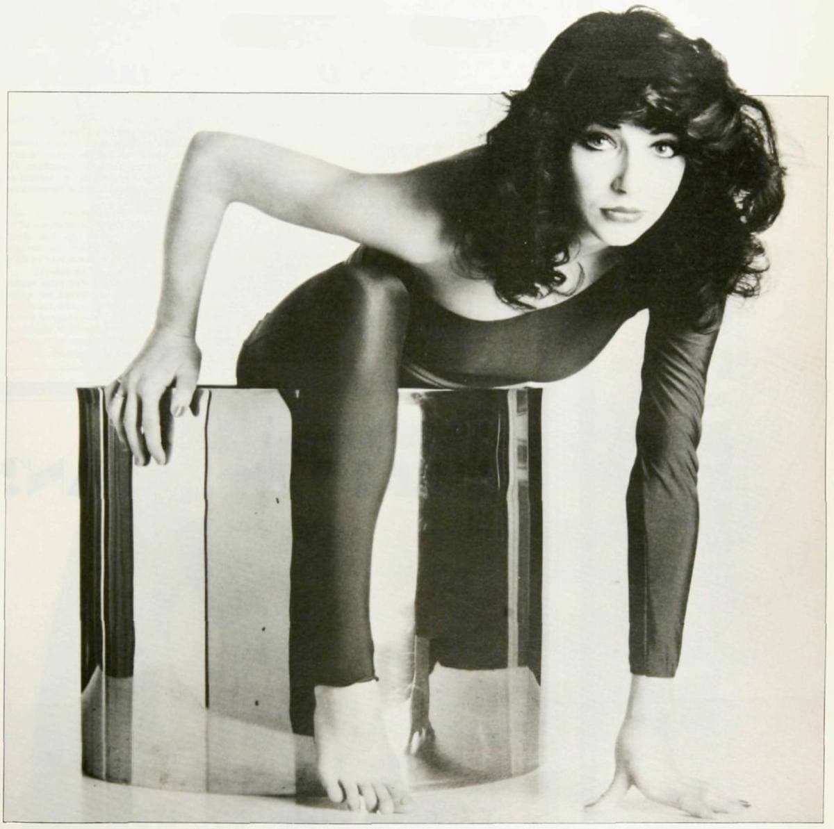 Kate Bush in a black body stocking, crouched on a clear perspex box, one arm reaching down, her hair loose