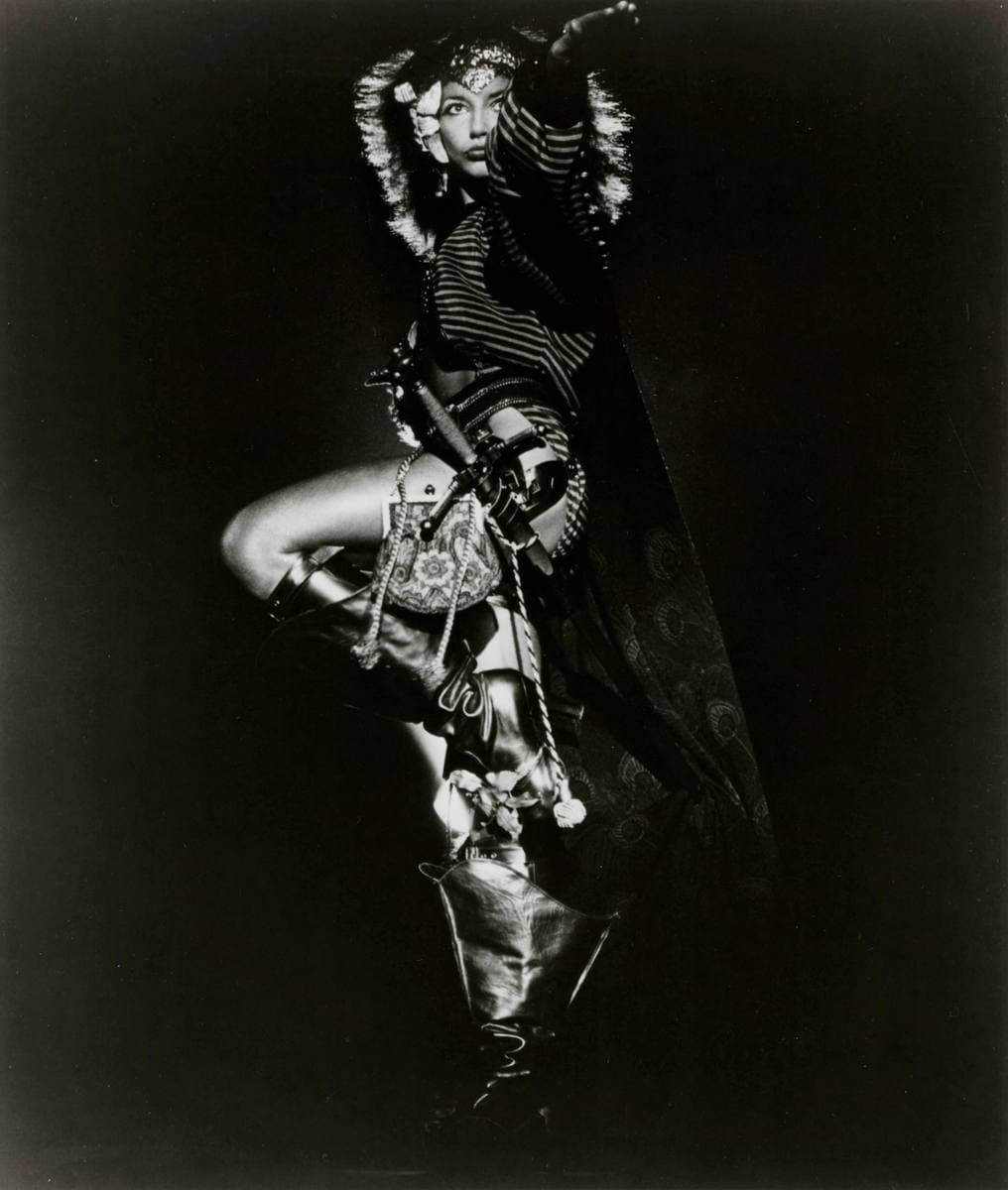 Kate Bush on a black background in a dark, patterned costume, one arm raised high above her head