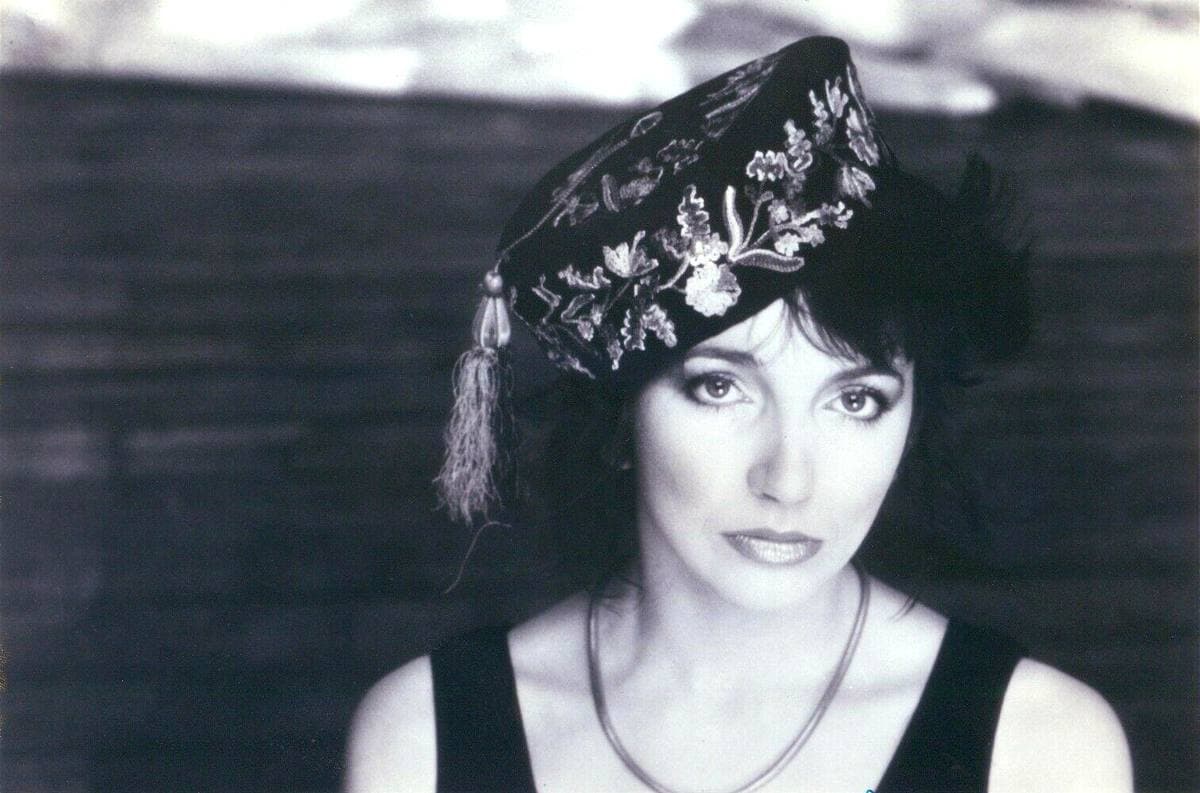 Kate Bush in black and white wearing an embroidered cap with a tassel and a black top, standing in front of dark water