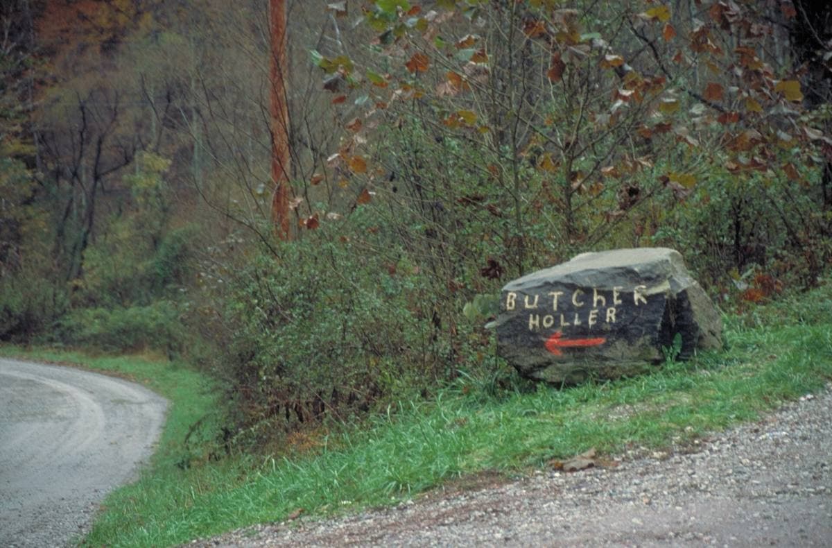 A grey painted boulder by a road with lettering pointing the way to Butcher Holler