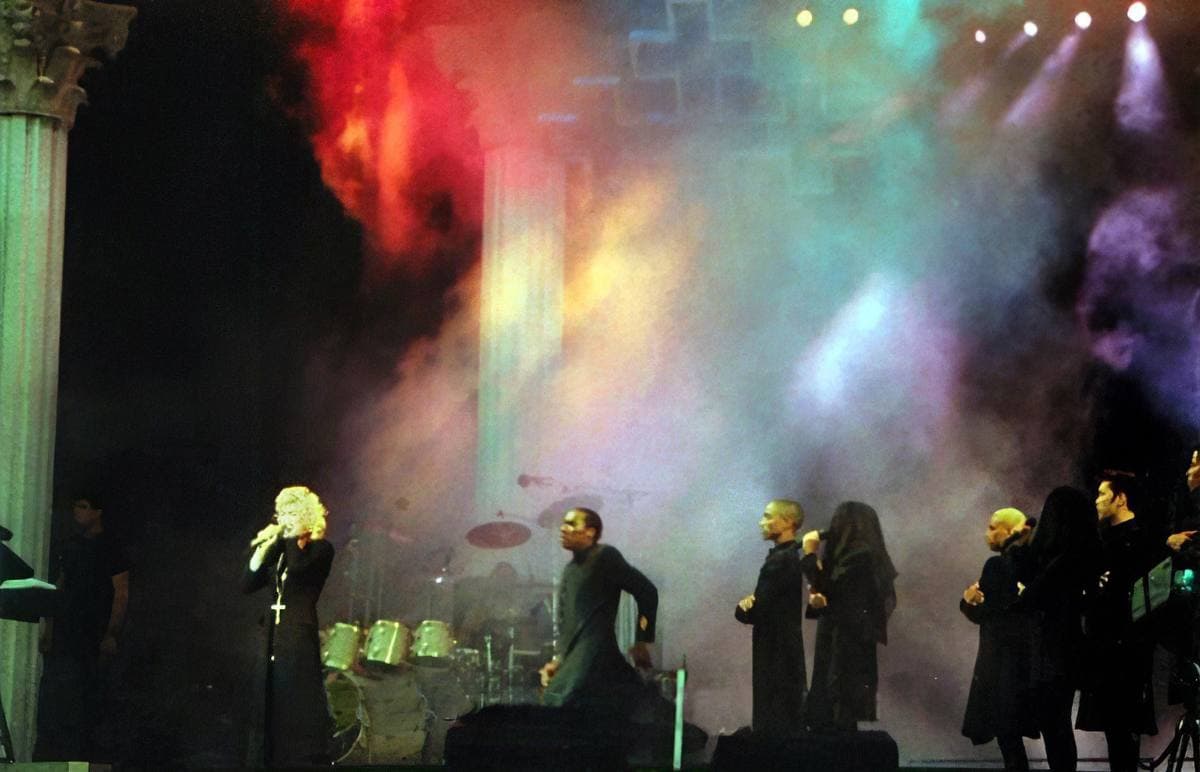 Madonna in a black cassock with a crucifix on a smoky stage of columns and coloured light, dancers in black robes beside her