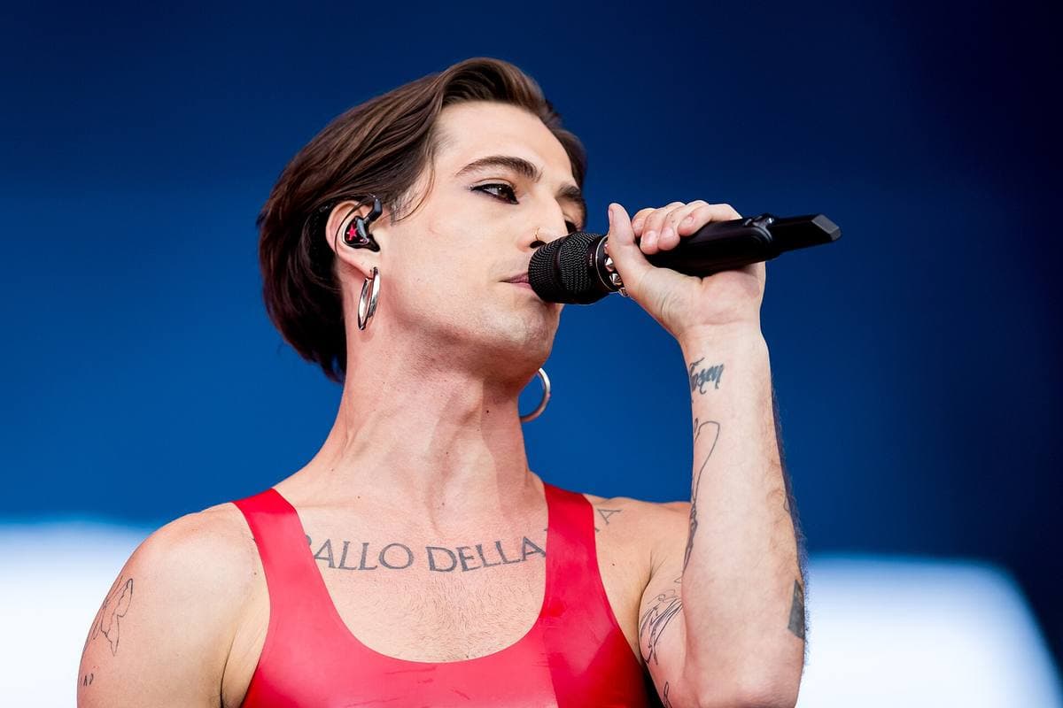Damiano David in a red vest singing into a microphone against a deep blue background