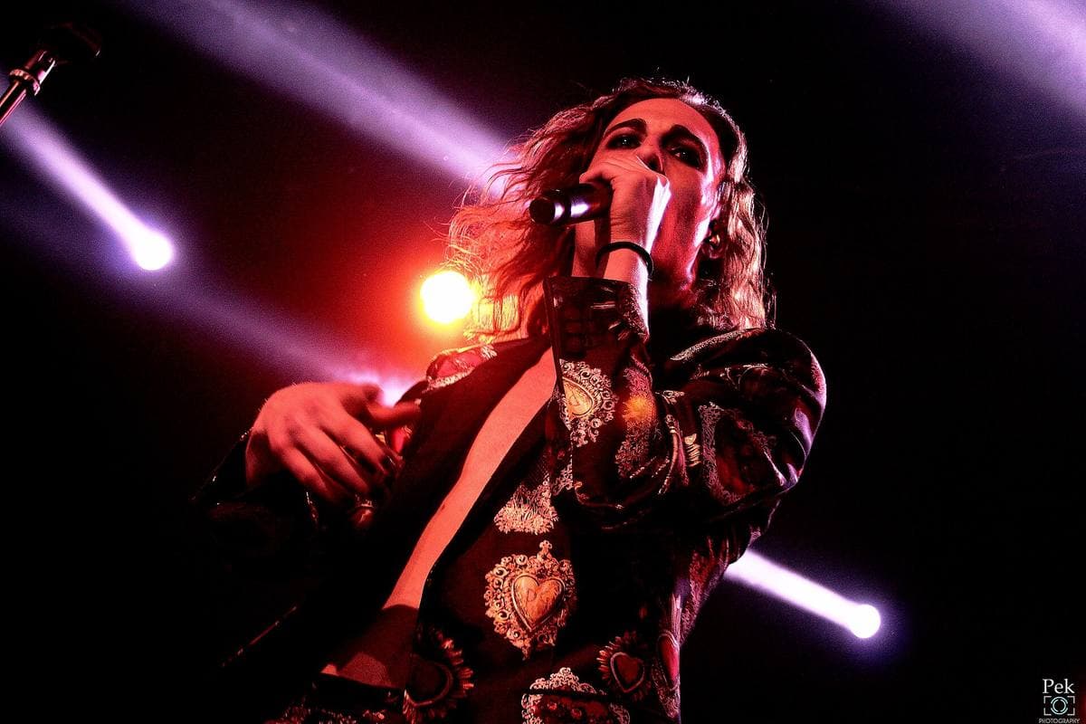 Damiano David singing into a microphone under purple and red stage light, in a patterned jacket