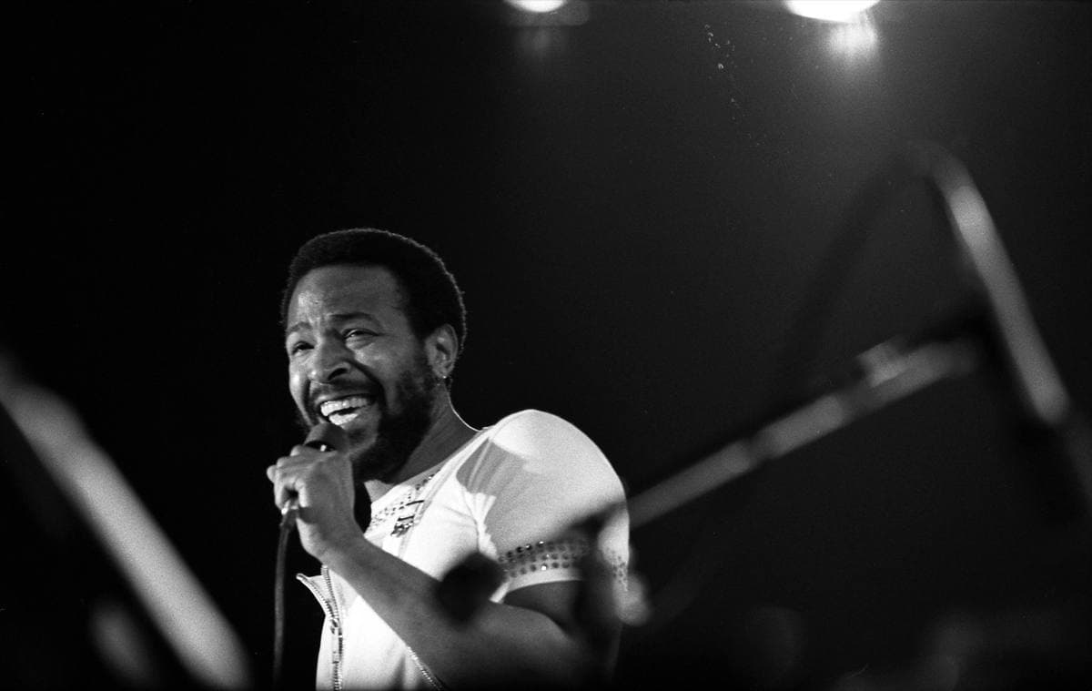 Marvin Gaye singing into a hand microphone under stage lights, in a white short-sleeved top
