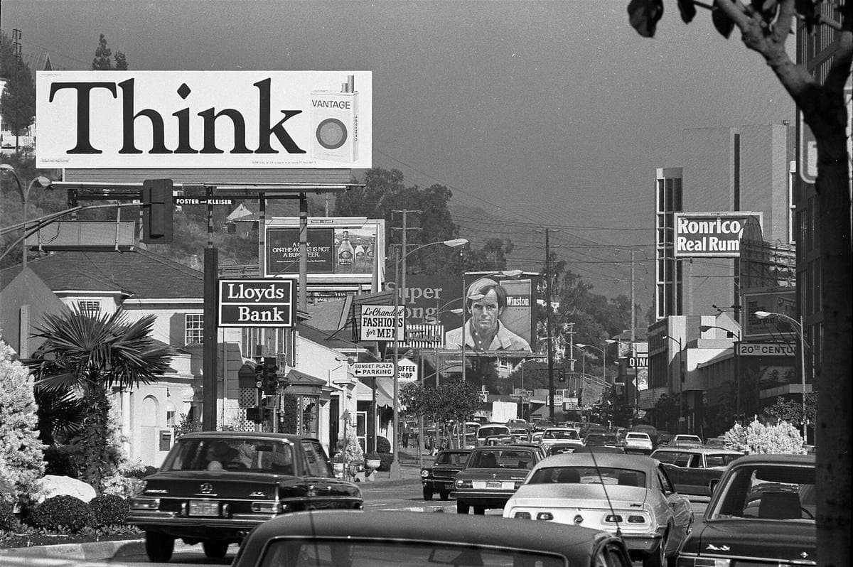 Black-and-white photograph of a busy boulevard lined with tall billboards and 1970s cars