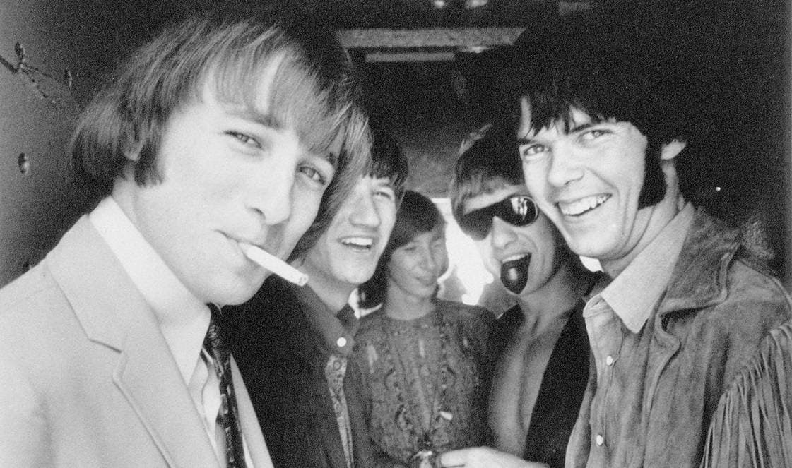 Black-and-white photograph of five young men crowded together and laughing; on the right Neil Young grins in a fringed suede jacket