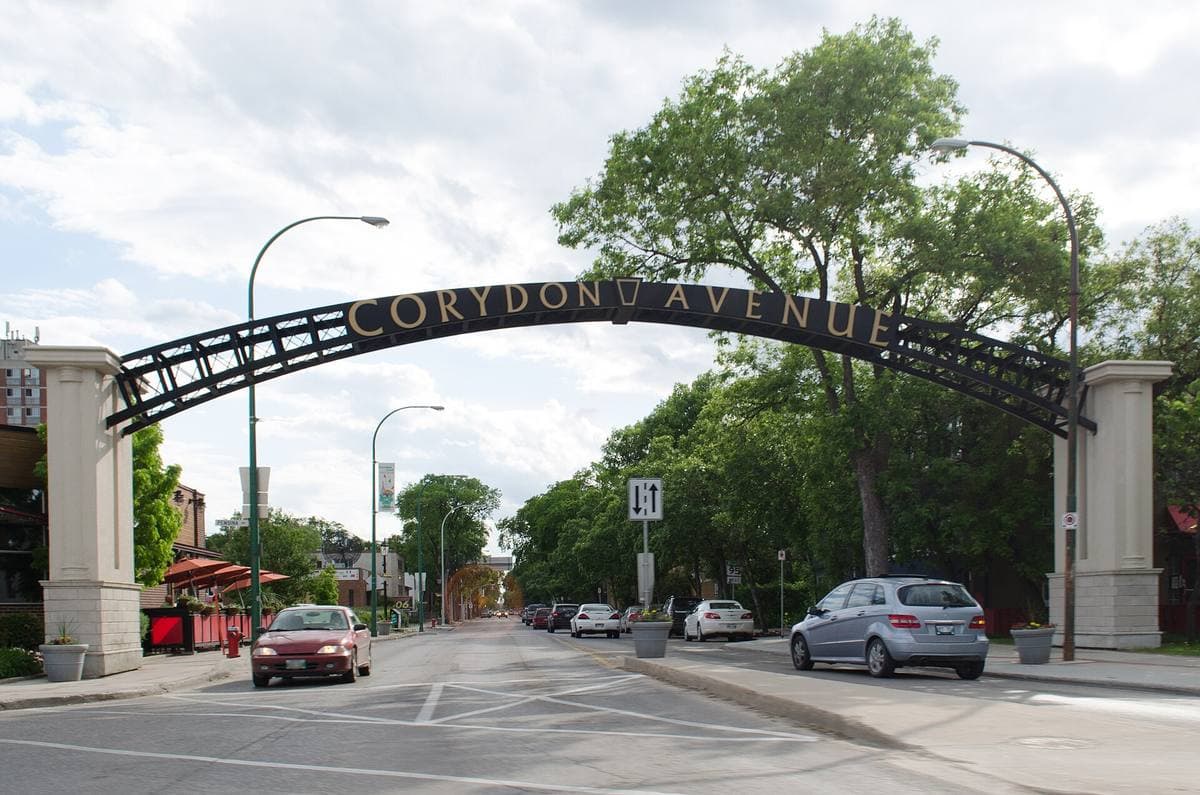 A black iron arch reading Corydon Avenue over a tree-lined street with parked cars