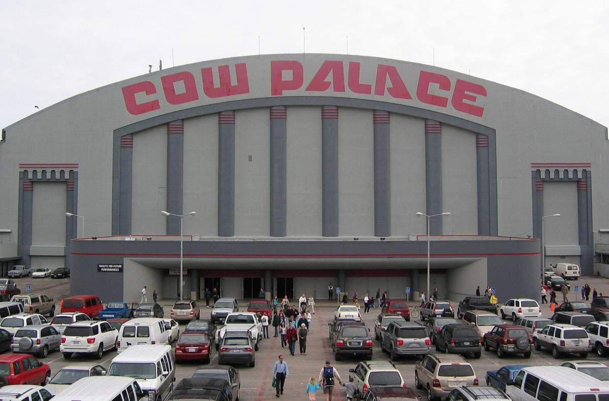 A wide arena front with Cow Palace in red letters over a curved roof, a car park in front
