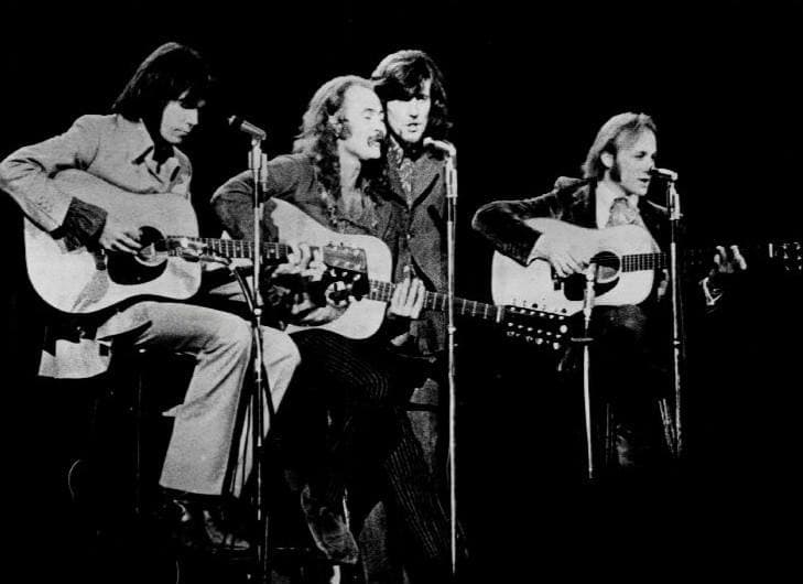 Black-and-white photograph of four men on stools with acoustic guitars at microphones; Neil Young on the left