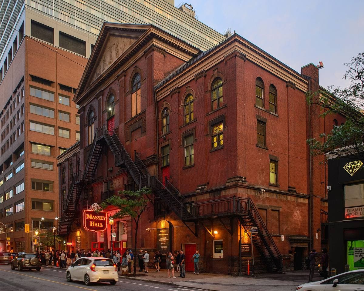 Red-brick concert hall at dusk with fire escapes and a lit canopy, towers behind