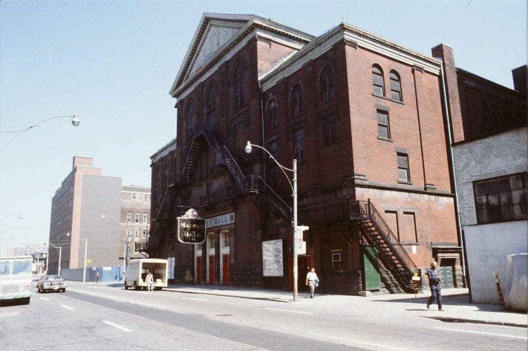 Colour photograph of a tall red-brick hall with iron fire escapes on its side, a quiet street in front