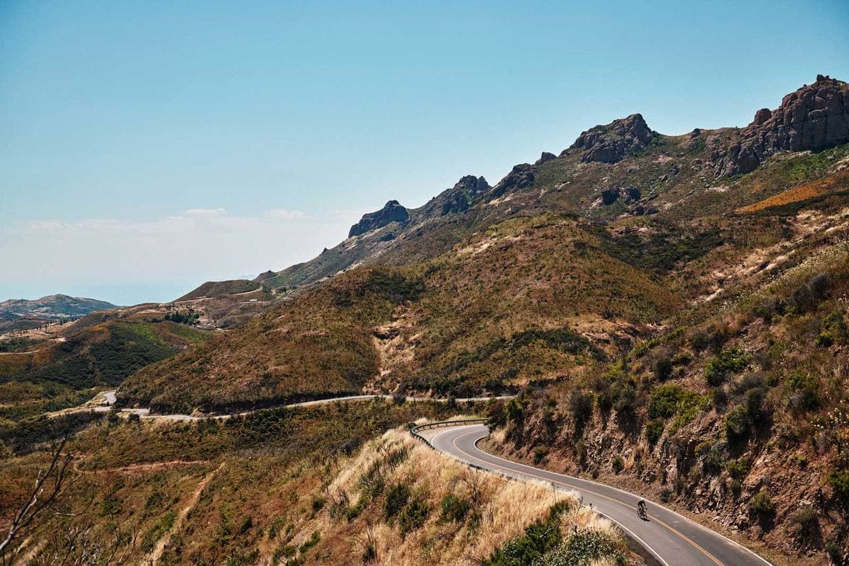 A winding road climbing through dry brown hills under rocky peaks