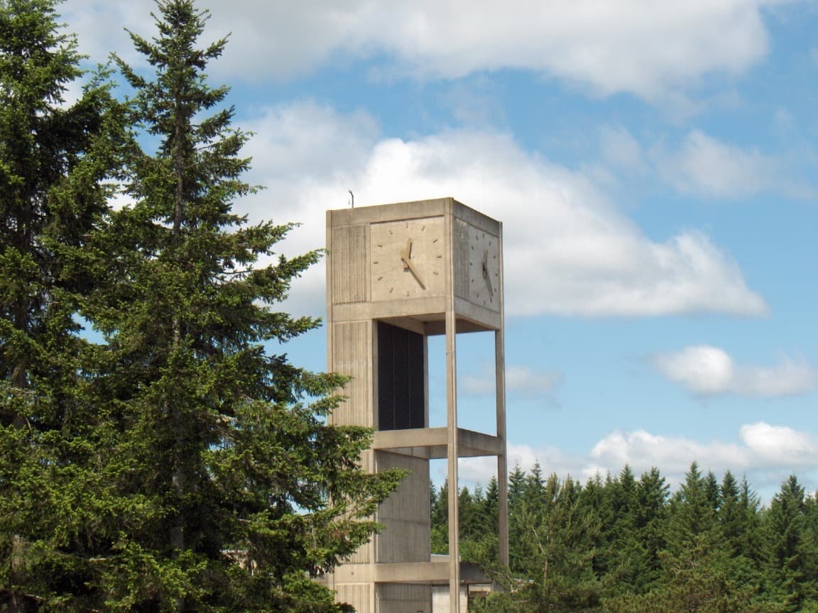 A concrete clock tower on the Evergreen campus among tall evergreen trees