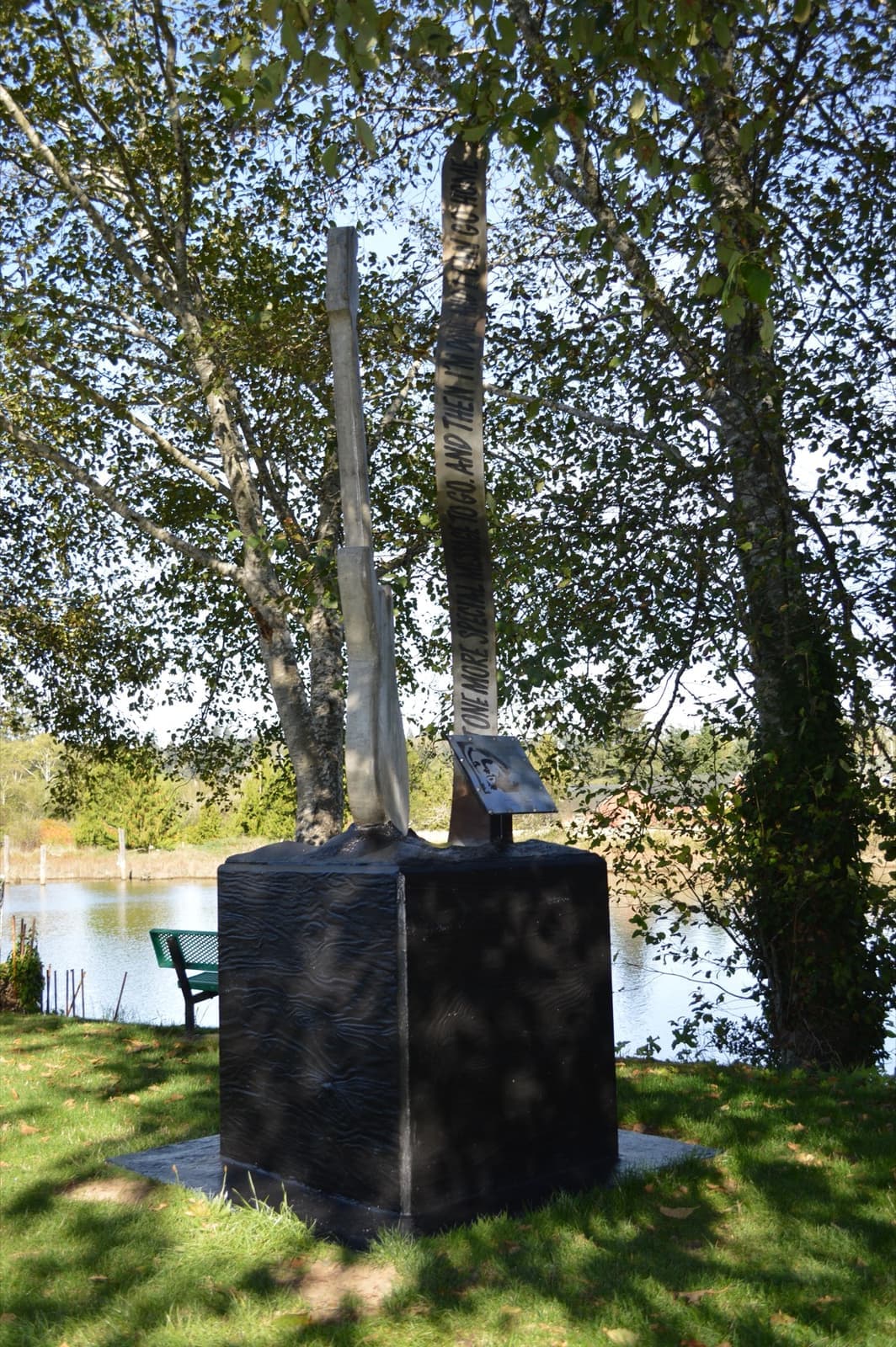 A tall pale concrete guitar and a steel ribbon on a black plinth, under birch trees by the river
