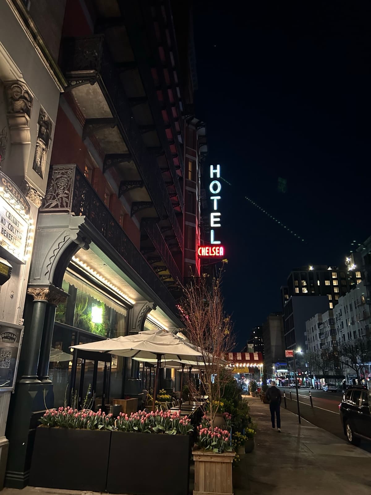 The red Hotel Chelsea sign glowing vertically at night above a pavement with planters and an awning