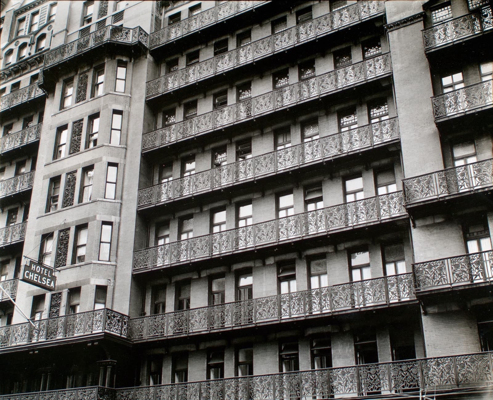 Black and white view looking up at rows of wrought-iron balconies on a tall brick hotel, with a small hotel sign