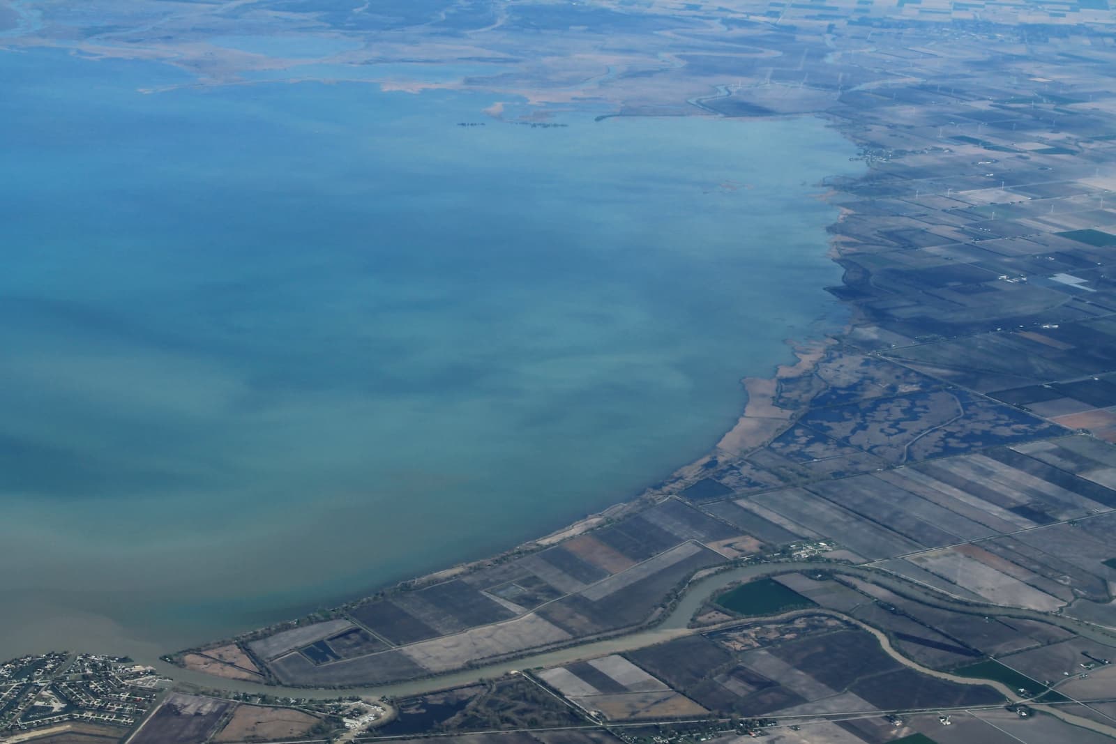 A wide blue-green lake meeting a flat shoreline of fields, seen from a plane