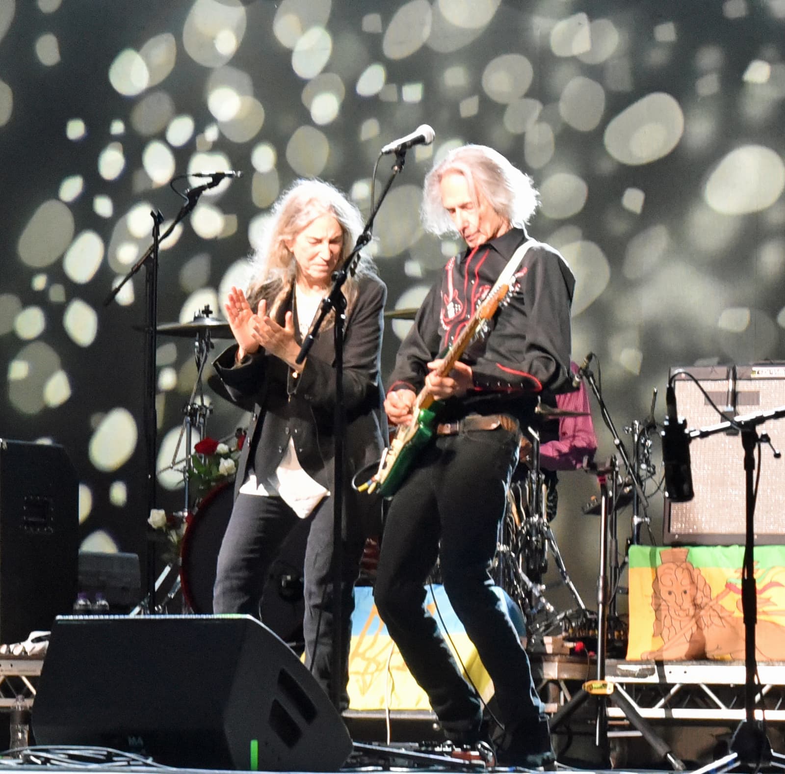 Patti Smith clapping beside Lenny Kaye playing a guitar, under spots of stage light