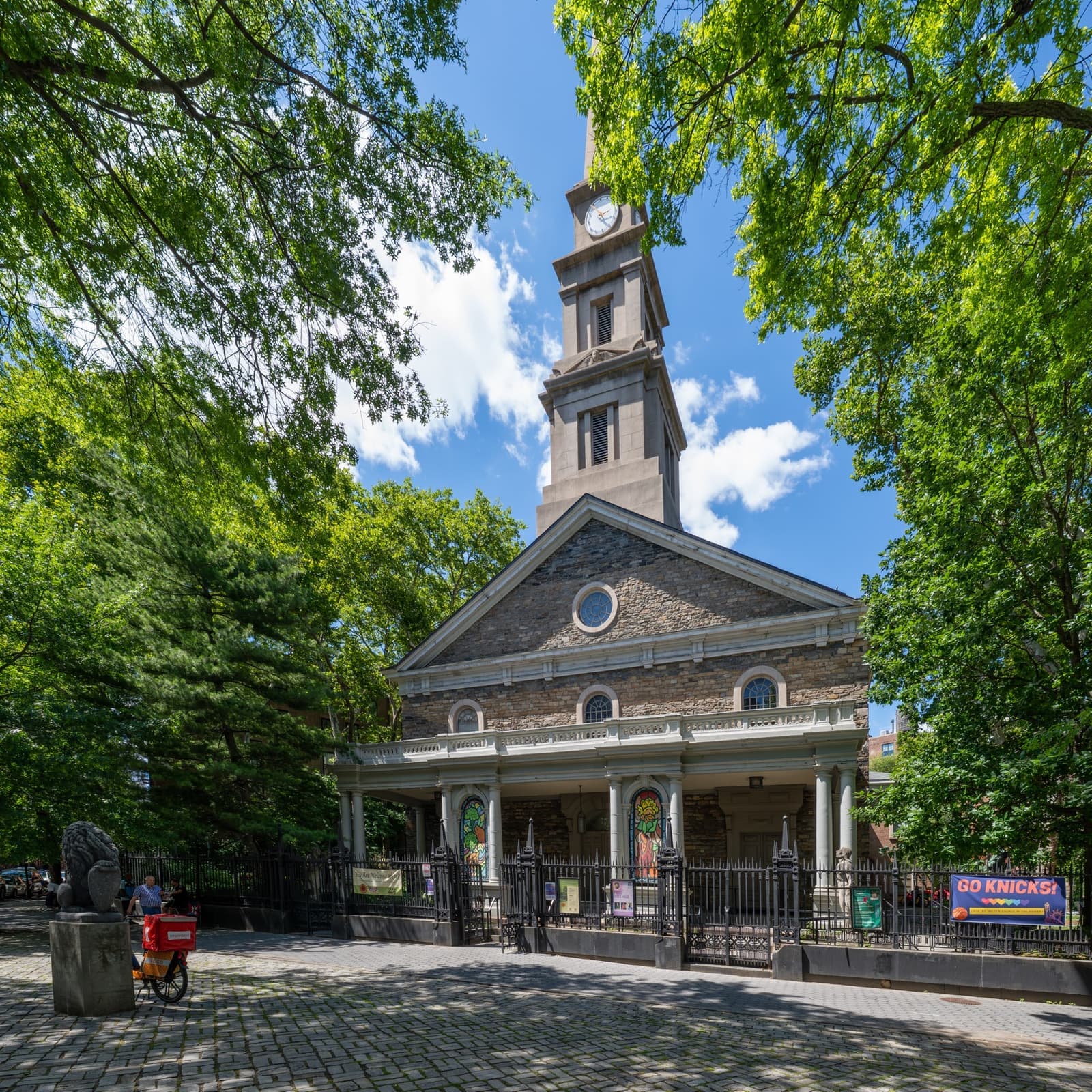 A stone church with a white steeple and a columned porch behind an iron fence and summer trees