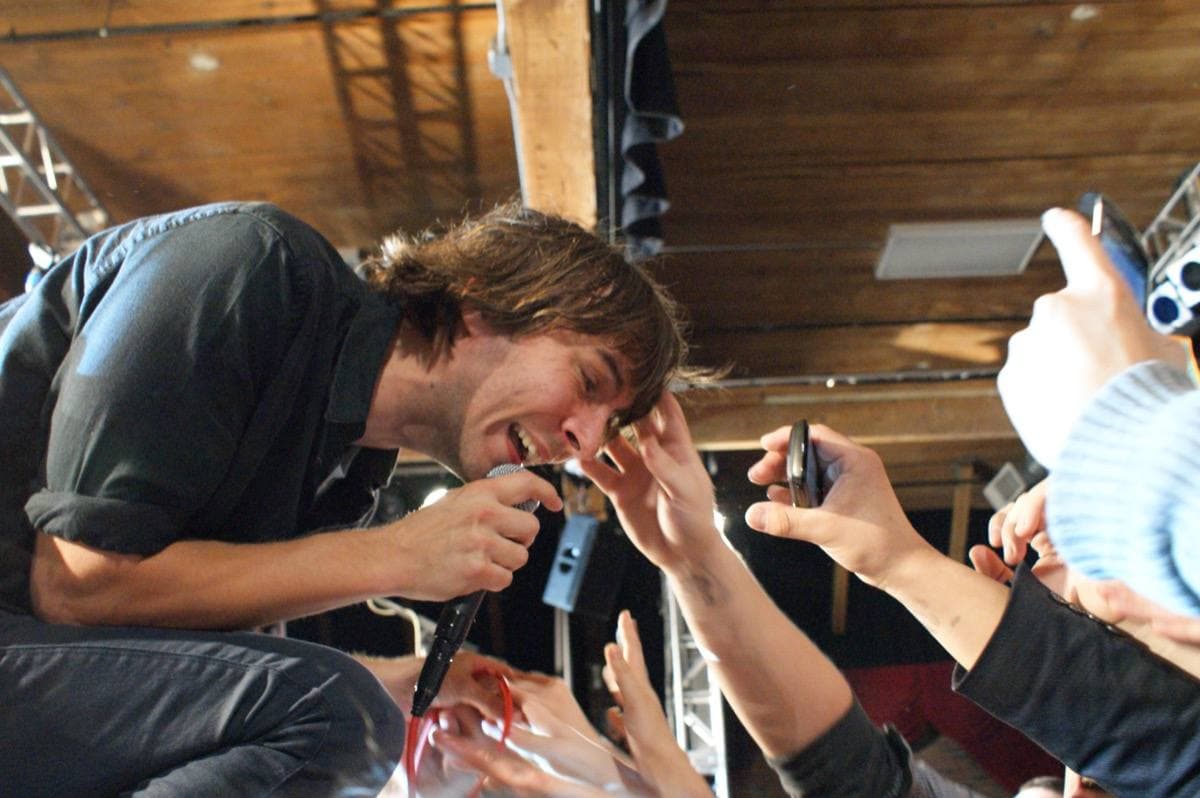 Thomas Mars bending down to the front row, fans reaching up, a wooden roof above