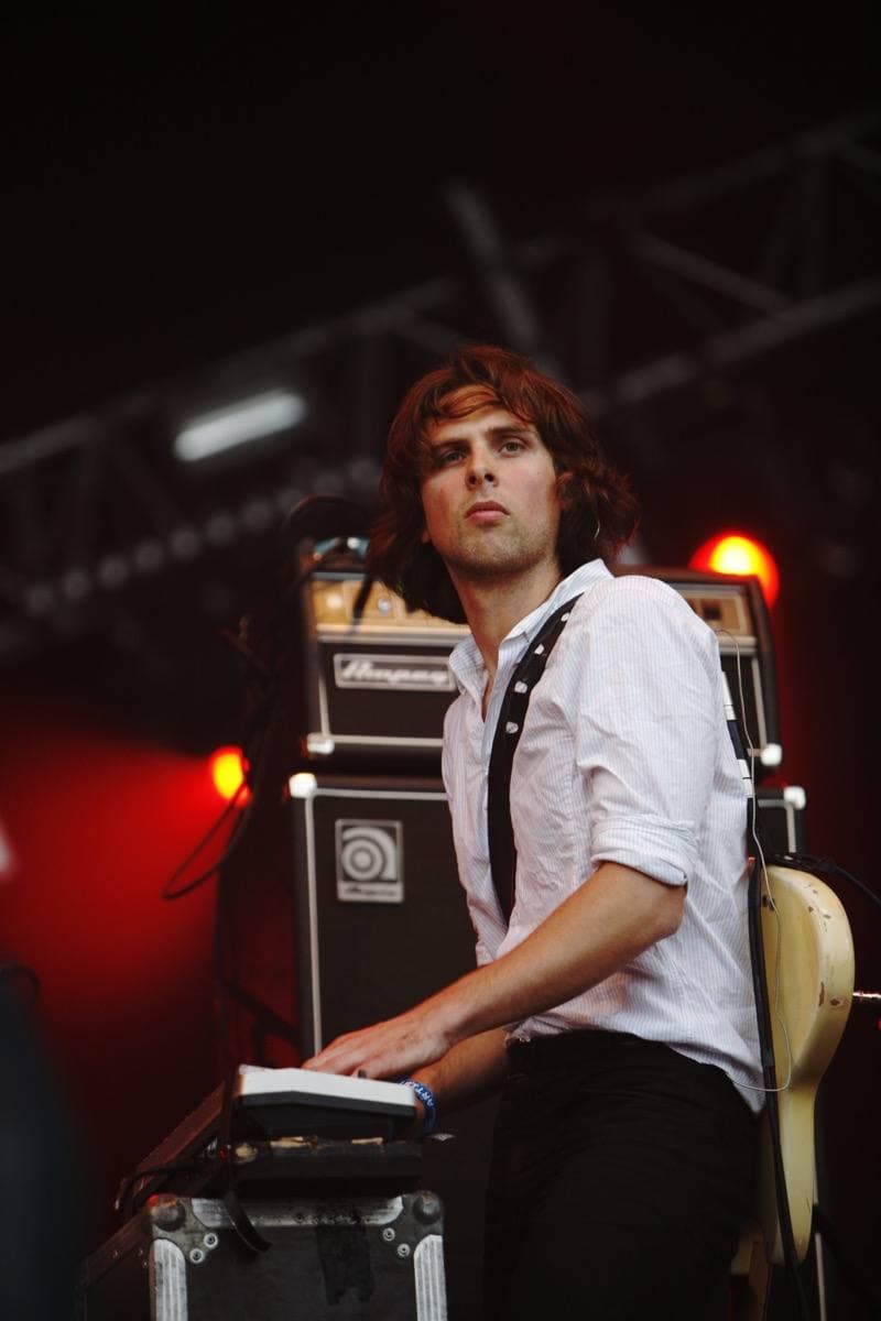 A long-haired musician in a white shirt at a keyboard, red light behind