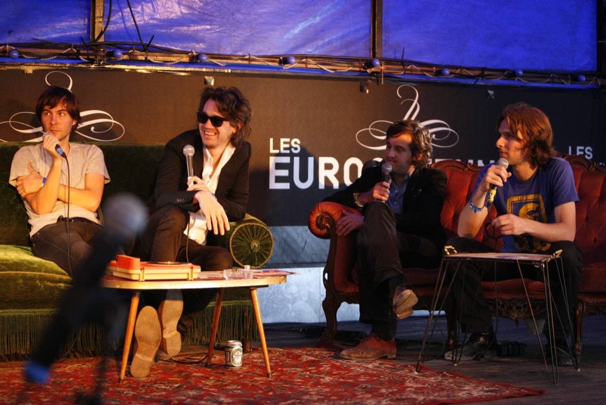 Young men on sofas and an armchair under a blue tent, talking into microphones